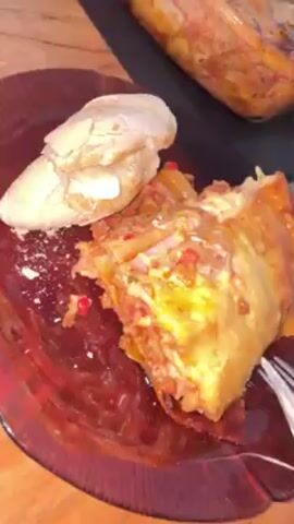 The video shows a close-up of a plate with a piece of lasagna and a small piece of bread with butter on top. A fork and knife are used to cut into the lasagna. The plate is red and has a unique shape. In the background, a glass baking dish with more lasagna is visible, with a serving fork inside. An orange and some small packets are also on the table. The table itself is a light brown wood grain.  The focus is on the food and the act of cutting and potentially eating the lasagna.