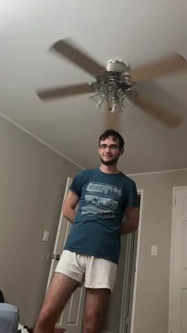 A young man with short, dark hair and glasses is featured in this vertical video, performing a sequence of gestures or a dance in what appears to be a bedroom. He is wearing a dark blue t-shirt with city skylines and the words "MADRID", "BERLIN", and "AMSTERDAM" printed on it, paired with light-colored shorts. The room has light grey walls, and a white door is visible in the background on the right. A spinning ceiling fan with three lights is prominently centered above the man. To the left, part of a bed with a blue blanket can be seen. The man's expressions range from a slight smile to having his tongue out, suggesting a playful or performative mood as he moves his hands in various patterns, including making a triangle shape with his fingers and gesturing with open palms. The lighting suggests it is daytime. The location is indoors, likely a residence in Franklin Township, United States of America.
