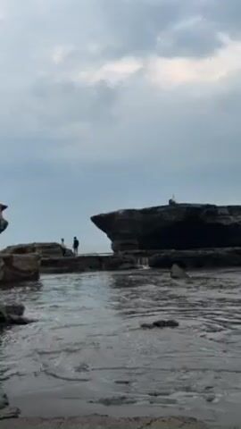 The video shows a rocky beach scene on a cloudy day. Two people stand on a rock formation close to a larger, flatter rock with something on top, possibly a small structure. Waves crash against the rocks. The tide appears to be low, revealing more of the rocky terrain. The water is dark and the sky is overcast. The overall impression is one of a moody, natural seascape.