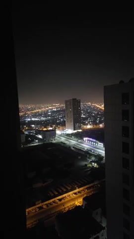A night time aerial view captures a city with numerous buildings and streets illuminated by lights. A tall skyscraper stands out in the center, with a parking lot visible in the foreground. The sky is dark, indicating nighttime.