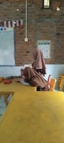 The video shows a classroom scene. In the foreground, a young boy wearing a light blue shirt is visible in profile. In the middle ground, a woman wearing a brown hijab and dress sits at a yellow table, looking down at something in front of her. A red object, possibly a book or notebook, is on the table. There is a green chair to the right of the table and a yellow chair to the right of that. In the background, several other people, including a woman in a blue hijab, are sitting on the floor. A whiteboard is mounted on a brick wall, with some writing and colorful circles on it. Decorative flags are hung across the top of the whiteboard. The room has tiled flooring and appears to be a classroom setting.