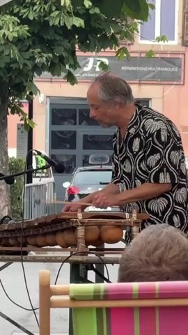 A man plays a balafon, a wooden xylophone with gourd resonators, outdoors. He wears a black and white patterned shirt and holds two mallets, striking the wooden bars. A microphone is positioned in front of the instrument. In the background, a building with a garage door is visible, along with trees and a car. Part of an audience member's head and shoulder is in the foreground, out of focus.