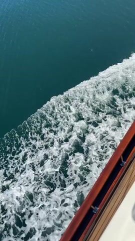 The video shows a close-up view from the side of a boat moving through water. The water is a deep turquoise color, and the boat leaves a foamy white wake. The side of the boat is visible, featuring a wooden railing and white exterior. The video is shot in daylight.