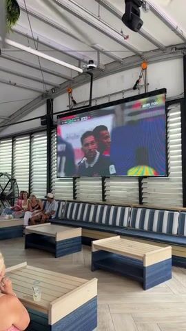 A wide shot captures an outdoor seating area with a large screen displaying a soccer match. Several people are seated on a long, striped couch facing the screen, with coffee tables in front of them. In the foreground, a blonde woman with long hair is seated, facing away from the camera. On the right side of the frame, a person in a pink shirt and turquoise shorts is tending to a coffee table. The environment appears to be a covered patio with overhead lighting and exposed ceiling structures. The video pans across the scene, showing different angles of the seating area and the screen.