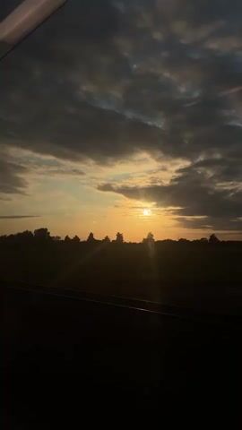 The video displays a sunset scene viewed from what appears to be a moving vehicle, evidenced by a visible window frame at the top. The sky is filled with dark grey and purple-tinged clouds across the top third, transitioning to a bright orange and yellow glow around the setting sun. The sun is visible as a distinct, bright circle, low on the horizon, partially obscured by the silhouettes of distant trees and fields that form a dark band across the middle of the frame. Sunlight radiates outwards from the sun, creating bright streaks. The immediate foreground is dark and indistinct, likely the interior of the vehicle or a dark part of the landscape passing by. Briefly, a tall, slender pole appears on the left side of the frame as the perspective subtly shifts.
