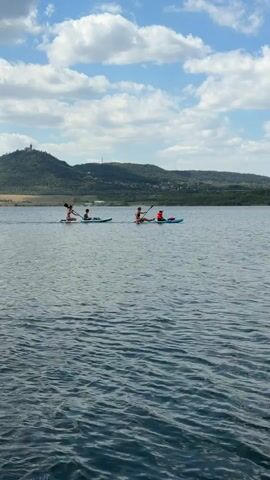 A wide shot captures a serene lake with a mountainous landscape in the background under a partly cloudy sky. In the foreground, the water is rippled and blue. Two kayaks are visible in the mid-ground, each carrying two people. The kayak on the left has two individuals seated, and the one on the right has two individuals seated, with one wearing a bright orange life vest. Both groups are paddling their kayaks across the water. The mountains in the background are green and rolling, with a small structure resembling a tower or castle visible on the highest peak. The overall impression is one of outdoor recreation and peaceful activity on a body of water.
