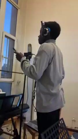 A young person is singing into a microphone. They are wearing headphones and holding a mobile phone. The individual is standing in a room with a window to their left, a keyboard, a laptop on a stool, and a music stand.