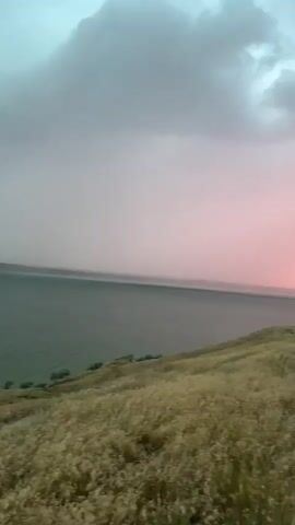 The video shows a landscape at sunset. There is a body of water on the left side of the frame, with a grassy hill in the foreground. The sky is filled with dramatic clouds that transition from gray and blue at the top to vibrant pink and orange near the horizon. As the video progresses, the camera pans to the right, revealing a dirt road, a utility pole with wires, and a few trees. In the distance, the outline of a building with solar panels and a car can be seen. The overall impression is a serene, natural scene with a striking sunset.