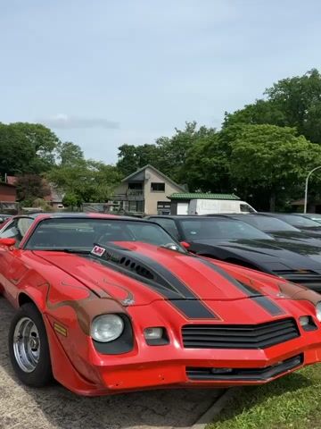 A red 1980s Chevrolet Camaro with flames painted on the hood and sides is parked outdoors. The car has a black racing stripe down the center of the hood and a large hood scoop. The sun is shining, and there are trees and buildings in the background. Another black car is parked behind the Camaro. The camera is positioned at eye level and appears to be moving slowly to the right.