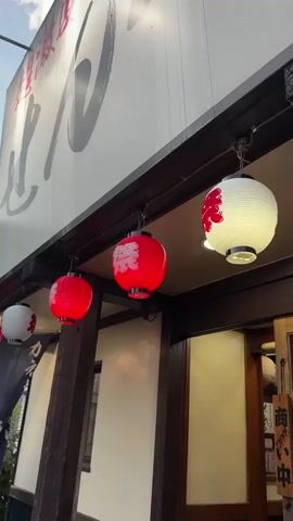 The video shows a row of four Japanese lanterns hanging from the eaves of a building. The lanterns are round and illuminated from within, casting a warm glow. Three of the lanterns are red with white Japanese characters, and one is white with red Japanese characters. A large white sign with black Japanese calligraphy is visible above the lanterns. The building appears to be made of wood and has traditional Japanese architecture. The lighting suggests it is daytime, but possibly overcast or late in the day as the lanterns are lit.