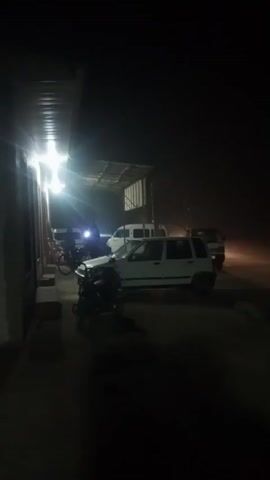 The video shows a white van driving by on a dark night. The van approaches from the distance, passing by the camera on the left and then continues down the road. The scene appears to be somewhere rural, with a building and parked cars visible in the background, lit by artificial lights. A motorcycle is also parked near the camera. The road and surrounding area are mostly dark, but the van's headlights and taillights are visible, and the streetlights illuminate the road. The video is taken from a stationary position.