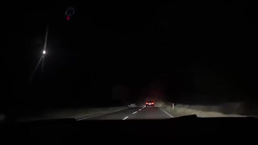 The video shows a point-of-view shot from inside a vehicle driving on a dark, winding road at night. The vehicle's headlights illuminate the road ahead, revealing a red car in the distance. There are trees and vegetation on either side of the road, and some small white markers line the edge of the road. A street lamp is visible on the left side of the road. There are also a few signs visible along the roadside. The overall impression is of a solitary drive through a rural area in the dark.