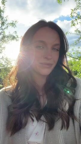 The video shows a woman looking directly at the camera, posing in front of a backdrop of trees and a partly cloudy sky. The sun is shining brightly behind her, creating lens flare. She has long, wavy brown hair, fair skin, and is wearing a light-colored cardigan over a white top with a floral pattern. At one point, she raises her hand to touch her hair. There are voices in the background.