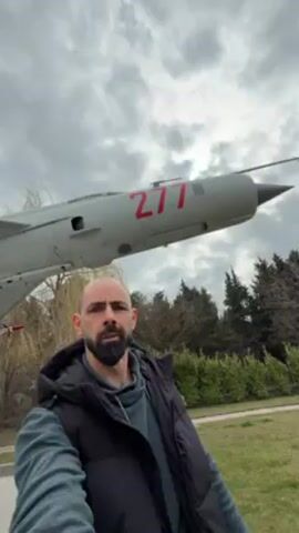 A man with a beard and wearing a black vest over a green top stands outdoors with a fighter jet on display behind him. The jet is light gray with the number 277 painted in red on its side. It is mounted on a concrete pedestal with a plaque. There are missiles attached to the wings of the plane. The sky is overcast, and trees are visible in the background. The man is looking at the camera and appears to be recording a video. The area around the monument is paved.