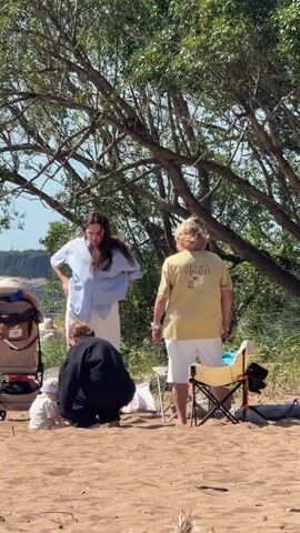 A group of people are gathered on a sandy beach under the shade of trees. A baby sits in the sand near a stroller. A person in a blue shirt and white pants stands and looks down, while another person in a black outfit bends over. A third person in a yellow t-shirt and white shorts stands facing away from the camera. A camping chair and bags are also visible on the sand. The background shows water and a distant shoreline with greenery.