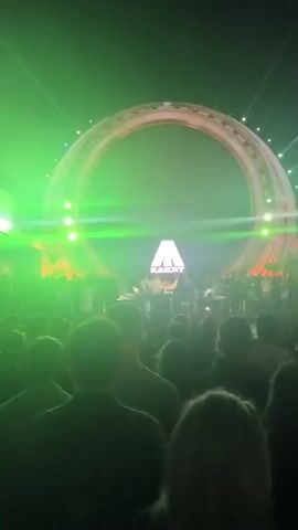 The video shows a concert or performance taking place at night. A large, circular stage structure with a dark center is visible, with a glowing logo in the middle. A crowd of people is gathered in front of the stage, with many of them holding up their phones to record. The stage is lit with various colors, including purple and red lights. Several performers are visible on the stage, some of whom appear to be DJs or musicians. The overall atmosphere appears to be energetic and festive.