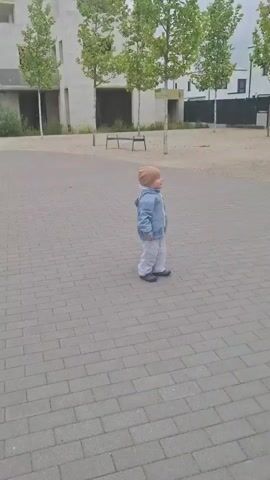 The video opens with a toddler standing on a paved walkway, facing right. They are wearing a light blue jacket, white pants, and a tan beanie. The background features modern white buildings with trees and benches. A man enters the frame from the right, holding a red and orange ball. He throws the ball towards the toddler, who turns to face it. The ball rolls towards the toddler. The toddler bends down and picks up the ball. The video ends with the toddler holding the ball.