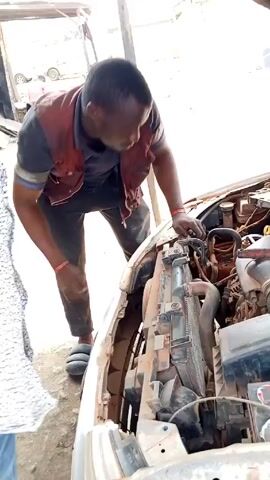 A man in a red vest is working on the engine of a car. The car's hood is open. The man is bent over the engine, appearing to be using a tool. He is wearing black pants and slides. The car is dusty and appears to be in a repair shop or outdoor garage setting. There are other people and vehicles in the background.