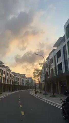 The video shows a street lined with modern, multi-story buildings, presumably in a residential or commercial area. The street curves slightly, with yellow lane markings in the center. The sky is visible above the buildings, showcasing a vibrant sunset with hues of orange and yellow peeking through the clouds. Street lights are positioned along the road, and there are sparsely-leaved trees planted along the sidewalk. A person on a motorcycle is seen on the right side of the road. The camera appears to be moving forward, potentially mounted on a vehicle.