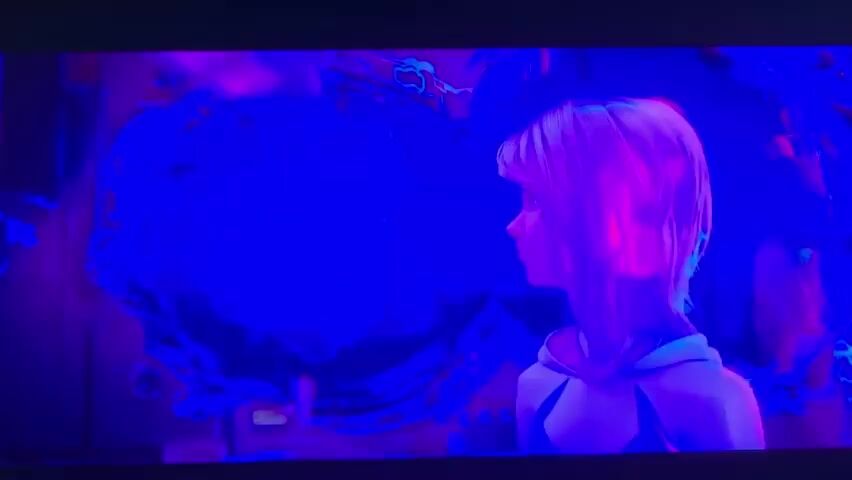 This is a video of a woman from the chest up, with bright pink and blonde hair and a dark top with a hood. She is looking to the right and her face is partially obscured by her hair, but she appears to be looking down. The background is a swirling vortex of blue and purple light, with some areas appearing darker than others. The lighting is dim and moody. There are some small, bright flashes of light that appear to be coming from the vortex. The woman's expression is unreadable but she appears to be serious or concerned.