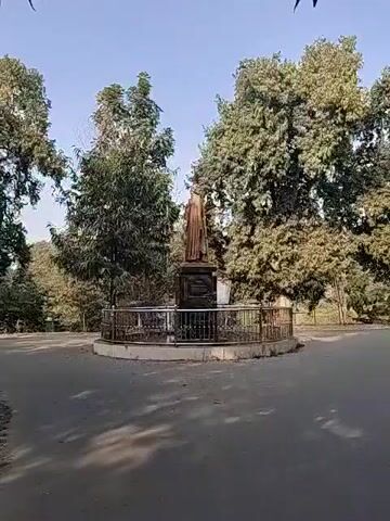 The video shows a bronze statue of a person standing on a stone pedestal in a park. The statue is enclosed by a low, metal railing. There are trees surrounding the statue and a paved area in front of it. The sky is blue.
