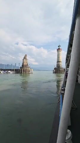 The video shows a view from a boat moving through a body of water, likely a lake or harbor. On the left, a stone statue of a lion stands atop a pedestal in the water, with a row of flags and moored boats visible behind it. To the right, a tall, cylindrical lighthouse with a red-capped top stands at the edge of the water. The water is a greenish-blue, and the sky is partly cloudy. The side of the boat, including a white railing and some ropes, is in the foreground on the right. A few people are visible on the deck of the boat in the distance. The scene appears to be a harbor entrance or a scenic waterway.