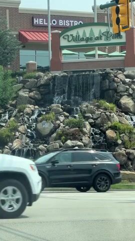 The video shows a street view in front of a shopping center called "Village at Pine" with a prominent waterfall feature. There are multiple cars driving on the street, some in motion and others blurred. The shopping center has a red awning and the words "HELLO BISTRO" are visible. The waterfall is made of rocks and water cascades down. The sky is visible and appears to be daytime.
