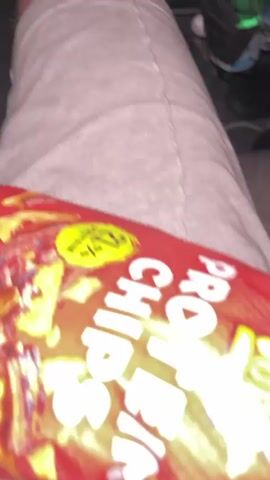The video shows a bag of "Rice Up" brand barbecue flavored chips. The bag is red and has images of chips and barbecue meat on it. The bag is resting on a person's lap. The person is wearing grey sweatpants. A blue cooler is visible in the background.