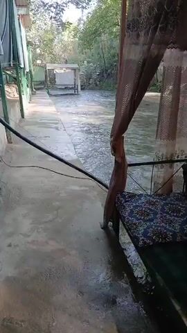 A slow pan reveals a narrow, concrete walkway next to a flowing river. On the left, a green awning covers a portion of the walkway. Further down the walkway, a white, open-sided structure with a sloped roof sits near the river. On the right, a wooden gazebo with a patterned cushion on a bench is partially visible. The river flows briskly, with ripples and small waves on its surface. Trees and buildings line the opposite bank of the river. The overall scene appears to be an outdoor seating area beside a natural watercourse.