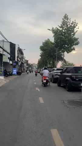 The video captures a street scene, likely in a Southeast Asian city, during what appears to be late afternoon or early evening. The perspective is from a moving vehicle, presumably a scooter or motorcycle, traveling down a paved road lined with buildings on the left and parked cars and palm trees on the right.

On the left side of the street, there are several low-rise buildings with storefronts. One sign is partially visible and appears to be in Vietnamese. Several motorcycles are parked along the sidewalk in front of these buildings. A few people are visible walking or standing near the shops.

The road itself has yellow dashed lines marking the lanes. Several motorcycles are in motion, traveling in the same direction as the camera.

On the right side of the road, a line of cars is parked. These include SUVs and what appears to be a van. The vehicles are parked along a raised curb or sidewalk area, beyond which are several tall palm trees and other greenery. A person on a scooter with a large red insulated bag is stopped near the parked cars, possibly making a delivery.

The sky is overcast with some hints of lighter color, suggesting the sun may be setting or has recently risen. The overall atmosphere is that of a typical urban street with active traffic and commercial activity.