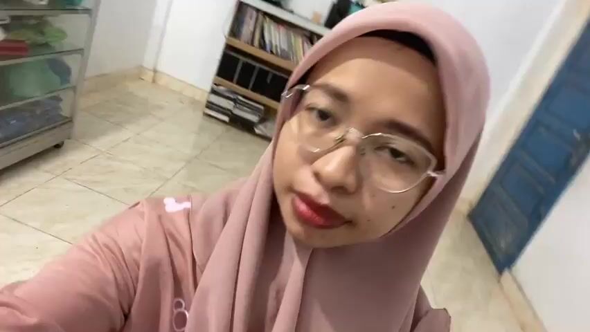 A young woman wearing glasses and a light pink hijab and a matching shirt is filming herself in what appears to be a room. In the background, there is a shelving unit filled with books and what looks like a slow cooker or rice cooker on top. To the left, there is a glass-fronted cabinet with items stored inside. To the right, a dark blue door is visible. The woman is touching her hijab and looking at the camera. The lighting is artificial and appears to be from overhead.