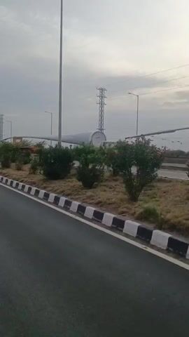 A wide shot shows a wind turbine blade being transported on a flatbed truck. The truck is driving on a road lined with bushes and a concrete divider. The sky is cloudy. The wind turbine blade is very long and white. There is a power line tower in the background. The truck is red and white and has a tarp over its cargo. The video pans to the right, showing more of the road and the surrounding landscape.