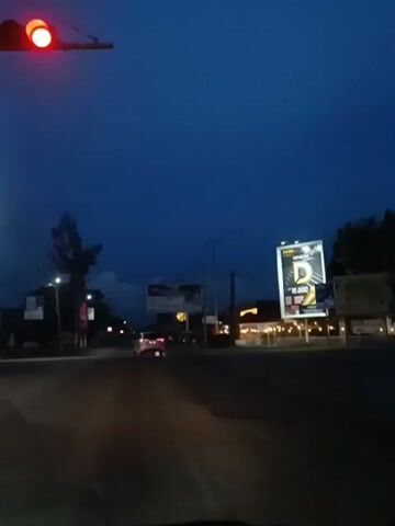 The video shows a street scene at dusk or night. A red traffic light is visible in the upper left corner. There are streetlights and illuminated billboards along the road. A few cars are visible on the road, some with their headlights on. The sky is dark blue. The overall impression is that of a street in a town or city during the evening hours.