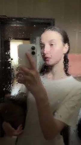A young woman takes a selfie in a mirror covered in water droplets. She is wearing a white t-shirt and her hair is pulled back. She is holding a white smartphone in her right hand and appears to be looking at the phone's screen. The background is blurred but suggests an indoor environment.