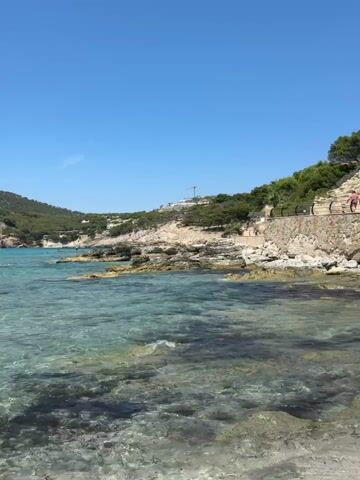 This media file depicts a scenic coastal view in Andratx, Spain. The scene is set on a bright, sunny day with a clear blue sky, suggesting it's midday or early afternoon. The turquoise water is crystal clear, allowing visibility of the rocky seabed below. Gentle waves lap against the shore, indicating calm weather conditions.

The foreground is dominated by the shallow, clear water with visible rocks and dark underwater vegetation. In the mid-ground, a rocky coastline curves around a bay. A stone wall with a railing runs along the top of the cliff, with a pathway behind it. Several people are visible enjoying the water and the beach. Some are swimming, while others appear to be wading or relaxing near the shore. One person is standing on the walkway at the top of the cliff, looking out at the sea.

In the background, lush green hills rise steeply from the coast, dotted with buildings. The presence of boats anchored in the bay suggests it's a popular spot for leisure activities. The overall mood is peaceful and idyllic, conveying a sense of a perfect summer day at the beach. No text is visible in the media file.