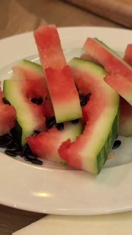 The media file shows a close-up of sliced watermelon on a white plate. The watermelon is cut into wedges and pieces, with some pieces appearing to have been eaten. Black seeds are visible scattered on the plate. In the background, another cutting board with more watermelon slices is visible, along with a fork. The setting appears to be a casual meal or gathering, possibly outdoors given the bright lighting. The overall mood is relaxed and summery. There is no visible text in the media file.