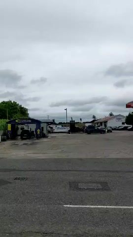 A panning shot shows a Texaco gas station under a cloudy sky. Several cars are parked at the pumps, and a Spar convenience store is attached to the station. A digital sign displays fuel prices. The camera moves from left to right, revealing more of the gas station and the surrounding area, which includes trees and other buildings. The video appears to be taken from a moving vehicle on a road.