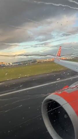 The video shows a view from an airplane window on a rainy day. The plane's engine, painted orange and red, is visible in the foreground. The wing of the plane, with the EasyJet logo on it, extends into the frame, pointing towards the right. Outside the window, a runway is visible, with other airplanes and airport buildings in the distance. The sky is cloudy, with hints of sunlight breaking through. Raindrops are visible on the airplane window, distorting the view slightly.
