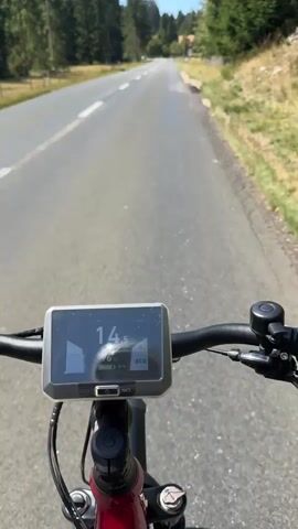 The video begins with a close-up view of an electric bicycle's handlebars and digital display, which reads "14.5" and "ECO." The bicycle is on a paved road that curves slightly to the left. Trees and vegetation line the right side of the road, while the left side features a white line marking the edge of the road. The camera is mounted on the handlebars, providing a first-person perspective of the ride. As the video progresses, the camera moves forward, showing more of the road ahead, which is surrounded by trees. The speed on the display changes to "15". The rider's legs are partially visible in the lower frame, and the shadow of the bicycle falls on the road. The scene is bright and appears to be a sunny day.