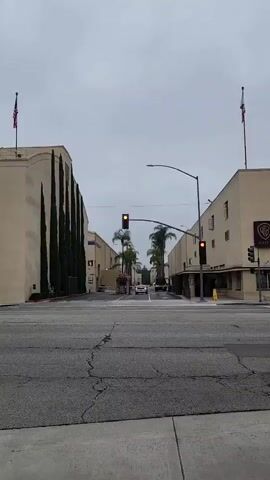 This media file shows a street view in Burbank, California. The scene is an intersection with traffic lights, palm trees, and beige buildings on either side of the road. On the left building, there are tall, dark green trees planted vertically, and an American flag is visible at the top. On the right building, a "WB" logo and the word "GATE" are visible, indicating this is likely a Warner Bros. studio lot.

Several cars are visible in the scene, including a white sedan and a black van. The traffic lights are red, indicating that traffic is stopped. The sky is overcast, suggesting a cloudy or potentially rainy day. The overall atmosphere appears calm and ordinary, depicting a typical day in a studio lot area. There are no people prominently featured in the frame, with the focus being on the architecture and vehicles. The cracked asphalt on the road adds a touch of realism to the urban setting.