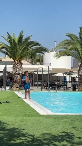 The media captures a lively day at a swimming pool in Cox, Spain. The scene is set under a clear blue sky, suggesting a warm and sunny day, likely in the late morning or early afternoon. Lush green artificial turf surrounds the vibrant blue water of the pool. Tall palm trees frame the poolside area, adding to the tropical atmosphere.

In the foreground, a man in black shorts and a white cap is in mid-action, leaning forward and appearing to be jumping or preparing to jump into the pool. To his left, a young child in a blue swimsuit stands watching. As the camera pans, the pool area reveals more activity. Several people are in the water, with one person making a large splash as they enter or move within the pool. Other individuals are seen relaxing near the poolside, some possibly sunbathing.

In the background, modern white buildings with balconies and awnings are visible, indicating a residential or resort-like setting. A black trash bin and metal railings are also present. The overall mood is one of enjoyment and recreation, with people engaging in typical summer pool activities. The bright sunlight casts distinct shadows, further highlighting the time of day. There is no visible text within the media.