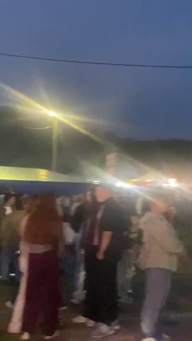 The video shows a crowd of people gathered outdoors at dusk. Bright lights illuminate the scene from various sources, including a street lamp in the upper left and what appear to be stage lights or carnival lights in the distance. The sky is dark blue, indicating it is evening. People are standing around, some in small groups, and there are tents or canopies set up in the background, suggesting an event like a festival or fair. The lighting creates lens flare effects in several parts of the video. Some individuals are looking in different directions, and there is a general sense of activity within the crowd.