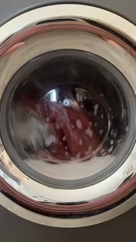 A washing machine is in operation. The inner drum is visible through a glass door, with clothes and soapy water swirling inside. The clothes appear to be red or dark colored. Bubbles of soap are visible on the surface of the water. The washing machine has a metallic rim around the glass door and a dark gray exterior. The lighting suggests an indoor environment.