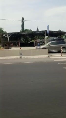 A car is seen driving past a crosswalk. A building with a metal roof is in the background. The sky is visible above the building.