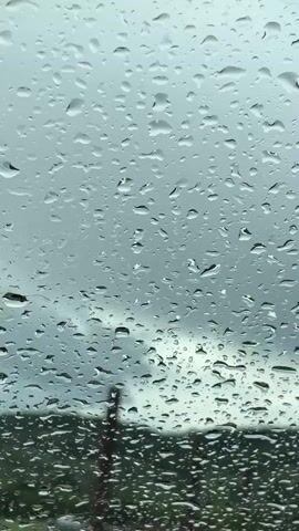 The video shows a car windshield covered in raindrops.  Outside the windshield, a blurred landscape of green hills and a cloudy sky can be seen. As the camera moves, the steering wheel and rearview mirror of the car come into view. There are no people visible in the video.