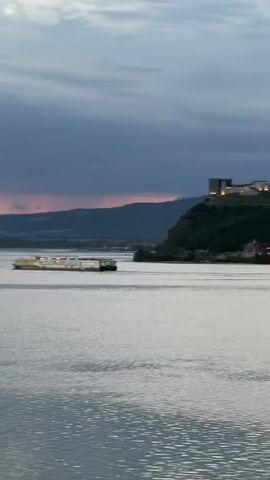 A drone shot pans over a large body of water. The sky is overcast and dusky, with hints of orange and pink from a setting sun peaking through dark clouds. In the middle ground, a long, white cruise ship travels from left to right. It has multiple decks and windows, with yellow accents along the top. Further to the right, on a hill overlooking the water, sits a building complex, lit up with lights. In the far distance, a mountain range forms the horizon. The water is calm and reflects the muted colors of the sky.