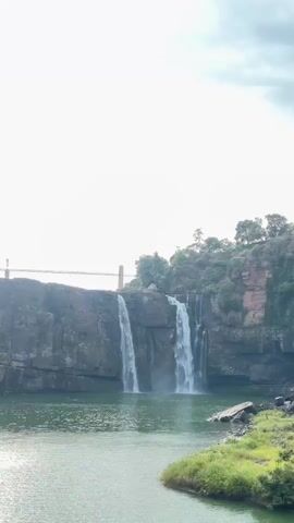 A vertical video showcases a waterfall cascading down a rocky cliff face into a body of tranquil, green water. The sun is visible in the bright, overcast sky. A bridge, supported by pillars, spans the cliff above the waterfall. Lush green vegetation covers the landscape, with trees visible on the cliff tops. The video pans out to reveal a wider view of the river, the surrounding cliffs, and the distant waterfall. There are no people visible in the video.