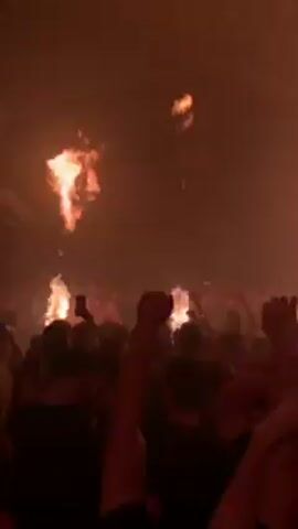 The video shows a crowd of people dancing at a concert. Initially, there are flames shooting up from the stage. Many people are holding up their phones, seemingly recording the event. The lighting shifts to a dark blue, with illuminated rectangular lights flashing above the crowd. The crowd continues to dance and record the event.