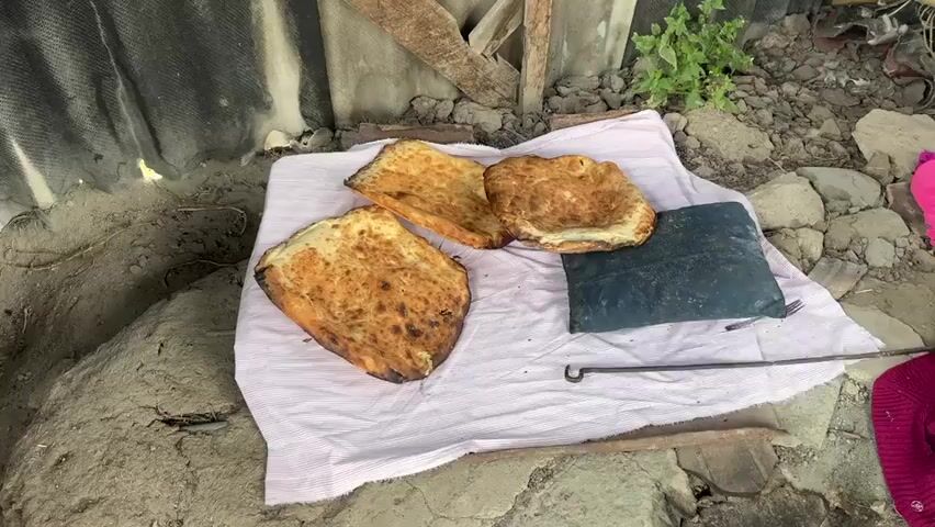 The video shows three flatbreads placed on a pink and white striped cloth. The bread is browned and appears to be baked. One bread is larger and positioned in the foreground, while two smaller breads are behind it. To the right of the breads is a dark rectangular object, possibly a cutting board or a mat. The cloth is laid out on the ground, which is a mixture of dirt, rocks, and what appears to be a rough, weathered surface behind the breads. The background includes some debris and a structure made of what looks like corrugated metal. There are no people visible in the video.
