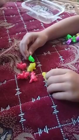 A child's hands are visible, interacting with small, colorful plastic dinosaur toys scattered on a red, quilted surface. The child is holding one green dinosaur in their right hand and another yellow dinosaur in their left hand. There are other dinosaur toys in pink and yellow colors arranged on the surface. In the background, a clear plastic container and some green and purple toys can be seen. The lighting suggests an indoor environment.